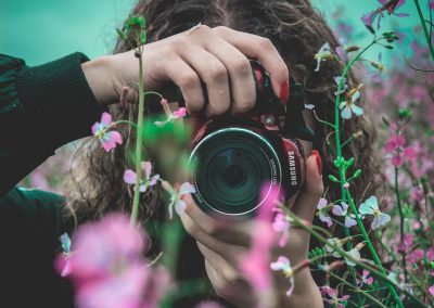 selective focus of woman behind pink flowers holding red Samsung bridge camera about to take photo of flower